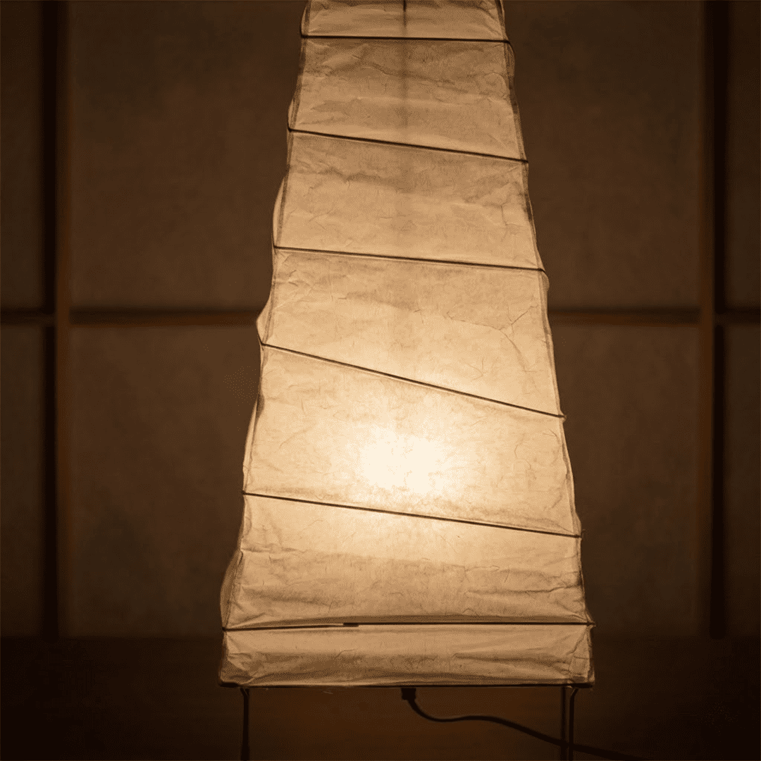 Handcrafted organic oval Washi Paper 4N Table Lamp on black tripod legs emitting a soft warm glow in a Japandi style interior setting.