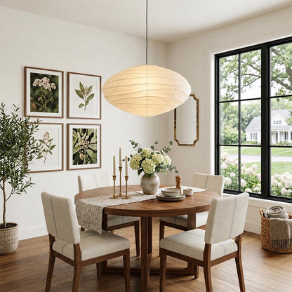 Minimalist Stuber Lighting oval rice paper pendant lamp glowing above a round wooden table, cozy Japandi style seating area with armchair and cane chairs.