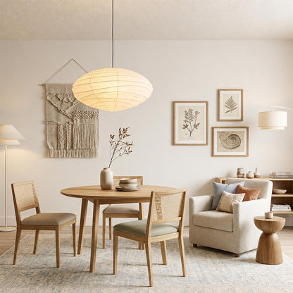 Minimalist Stuber Lighting oval rice paper pendant lamp glowing above a round wooden table, cozy Japandi style seating area with armchair and cane chairs.