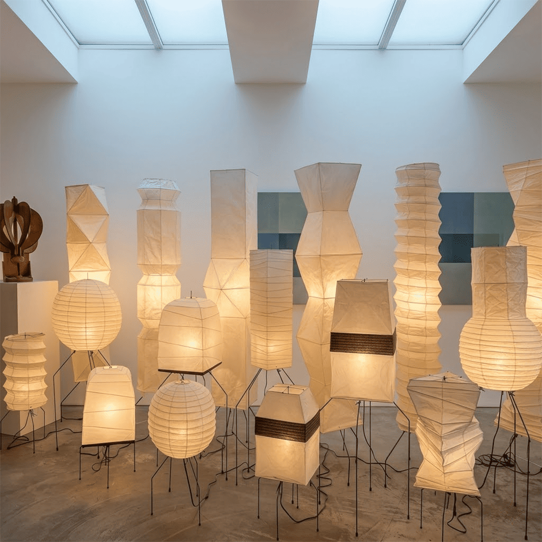 Large Akari UF3-DL floor lamp with a white washi paper shade on a black tripod metal stand, glowing with warm light in a minimalist room.