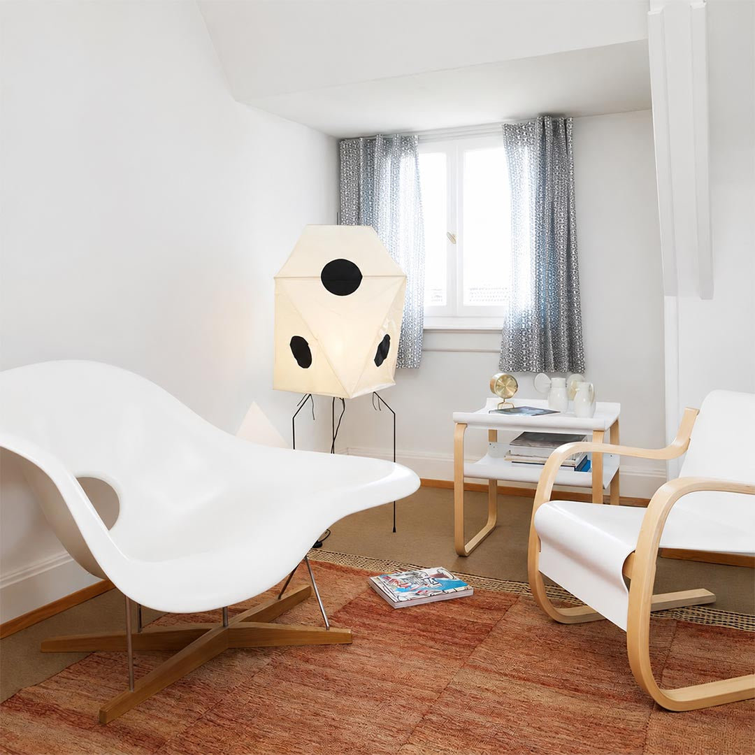 Mid-century modern origami floor lamp in a bright living room with white accent chairs and red rug.