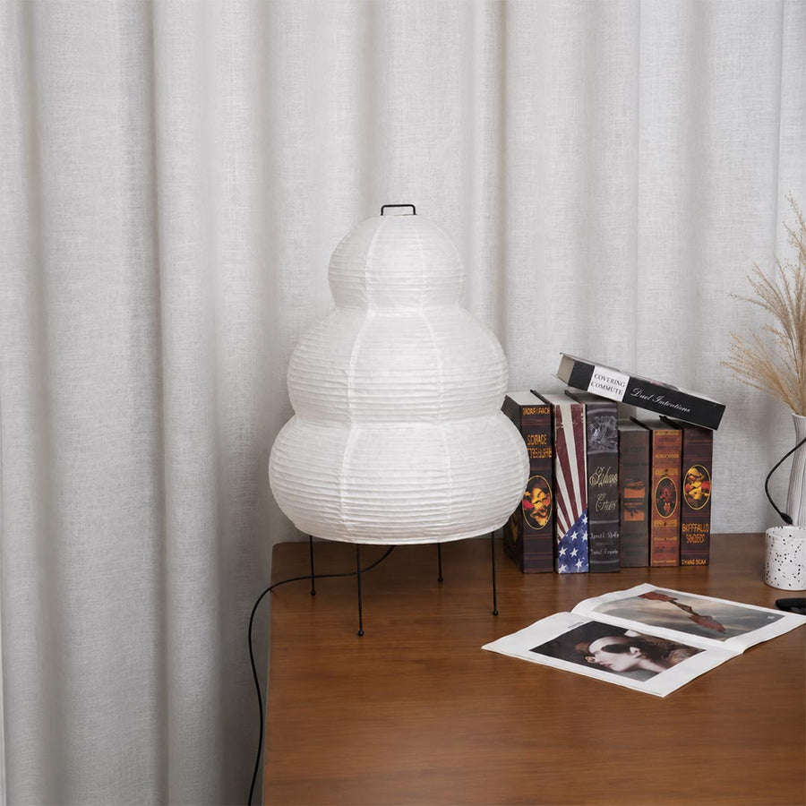 Unlit white washi paper lantern table lamp sitting on a wooden desk against white curtain backdrop