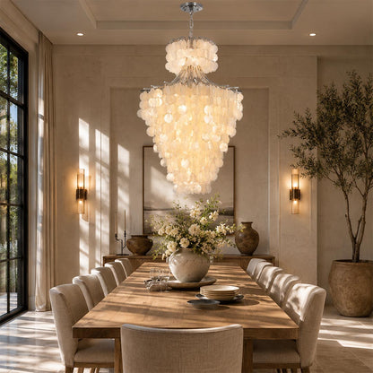 Minimalist statement lighting fixture over a long wooden dining table surrounded by beige chairs and warm wall sconces.