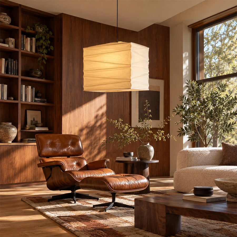Washi paper square cube pendant light hanging in a sunlit mid-century modern living room with a leather lounge chair and wooden bookshelves