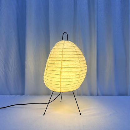 Washi paper tall tripod table lamp glowing warmly with soft ambient light against a cool blue curtain backdrop