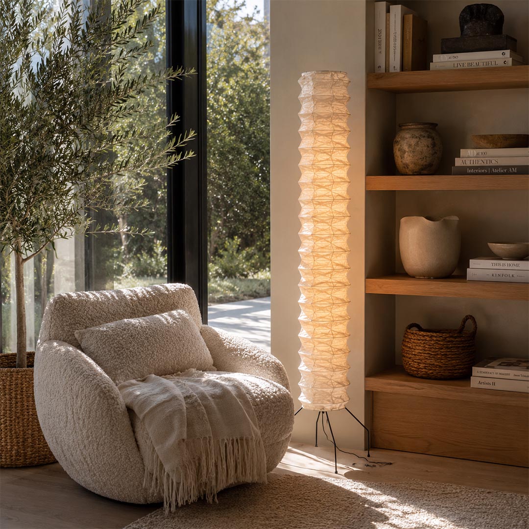 A cozy Japandi reading nook featuring a glowing Washi Paper UF4-31N Floor Lamp next to a plush lounge chair and wooden shelves by Stuber Lighting.