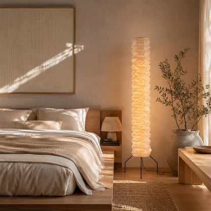 A cozy Japandi reading nook featuring a glowing Washi Paper UF4-31N Floor Lamp next to a plush lounge chair and wooden shelves by Stuber Lighting.